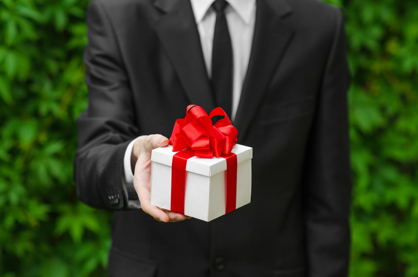 Gift and business theme: a man in a black suit holding a gift in a white box with a red ribbon on a background of green grass - Valokuva, kuva