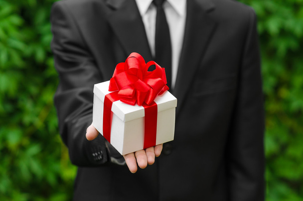 Δώρο και επαγγελματίες θέμα: ένας άνθρωπος σε ένα μαύρο κοστούμι κρατώντας ένα δώρο σε ένα λευκό κουτί με μια κόκκινη κορδέλα σε φόντο πράσινο γρασίδι - Φωτογραφία, εικόνα