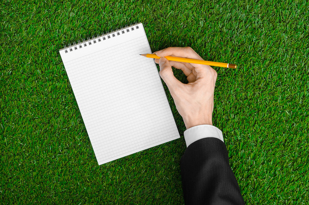 Day of knowledge and business topic: the hand of man in a black suit holding a notebook and pencil top view on a background of green grass - 写真・画像