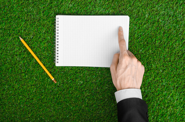 Day of knowledge and business topic: the hand of man in a black suit holding a notebook and pencil top view on a background of green grass - Φωτογραφία, εικόνα