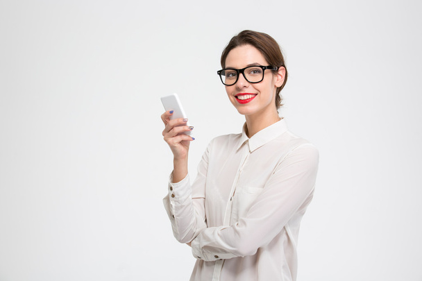 Cheerful attractive businesswoman in glasses standing and using mobile phone - 写真・画像
