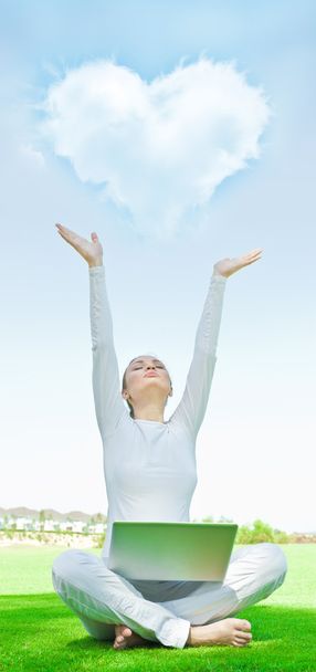 Довольно расслабленная женщина, сидящая на траве и вытягивая руки, прицеливается в небо, разговаривая на ноутбуке. Облачное сердце над головой
 - Фото, изображение