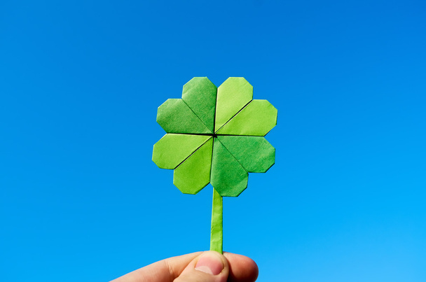 Hand holding green paper origami folded shamrock on blue sky background. Sunny weather outdoors. - Foto, afbeelding