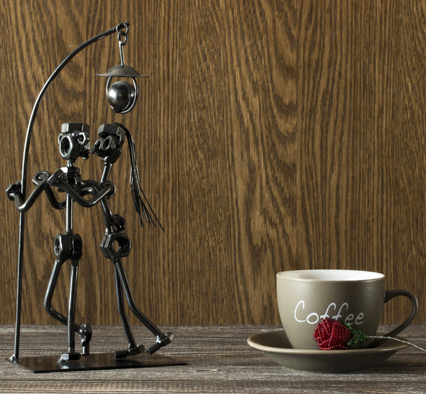 Статуэтка влюбленной пары, целующейся под лампой из металлических винтов и стержней рядом с чашкой кофе и искусственными бусами из роз
 - Фото, изображение