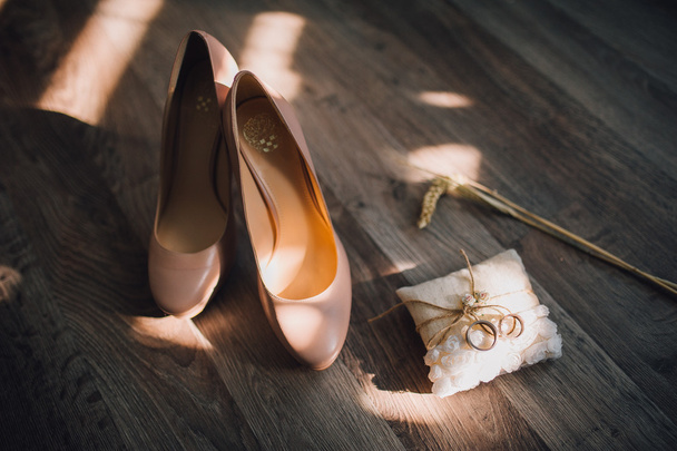 beige bridal shoes laying on wooden floor with ears of wheat and pillow with wedding rings on side - Photo, Image