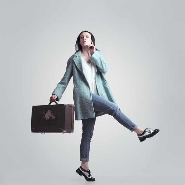 Young brunette girl with a suitcase. Isolated on a gray background. To go wide step. - Fotografie, Obrázek