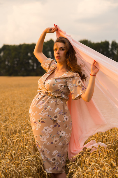 Pregnant woman with a handkerchief in his hands in a wheat field. Happy woman in a field. A woman enjoys the unborn child. Sincere emotions and joyful feeling. Beautiful countryside. Wheat field awaiting harvest. - Fotografie, Obrázek